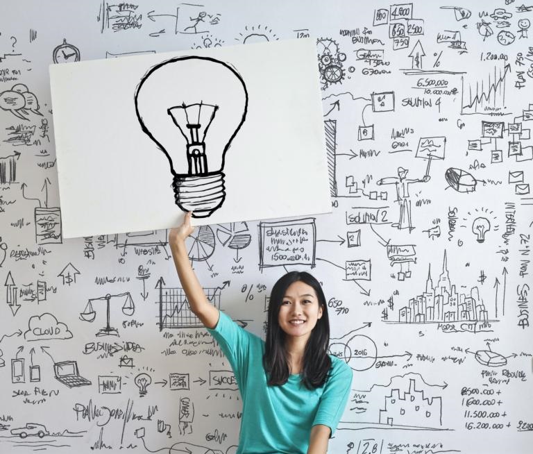 Dame sitzt vor bemalten Whiteboard und hält Plakat mit gezeichneter Glühbirne in der Hand